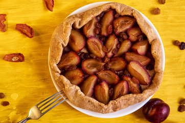 Plums pie with raisins on a white plate on yellow wooden background decorated with fresh plums, brown raisins, dried pieces of apple and silver fork. Top view