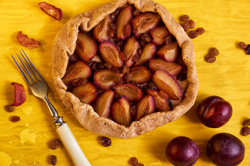 Homemade plums pie with raisins on yellow wooden background decorated with three fresh plums, brown raisins, dried pieces of apple and silver vintage fork. Top view