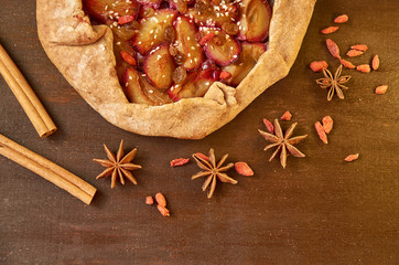 A half of plums pie with raisins, goji berries and sesame on brown wooden background. Pie with plums decorated with dried goji berries, anise stars and cinnamon sticks with copy space. Top view
