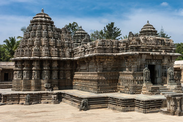 Mysore, India - October 27, 2013: Beige stone central sanctuary, called Trikuta, of Chennakesava Temple in Somanathpur under bluish sky. Fully decorated with statues and friezes. Tops of green palms.