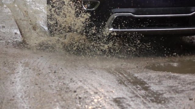 SLOW MOTION CLOSE UP: Detail of a SUV car tyre driving over deep puddle and splashing water across the dirt road. Black SUV car tires driving on wet dirt road after rain and over muddy puddle.