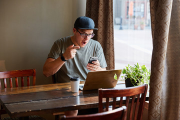 man on smart phone excited next to computer at coffee shop