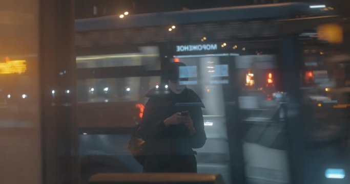 Woman using mobile while waiting at bus stop at night. Car traffic in background, view through the glass
