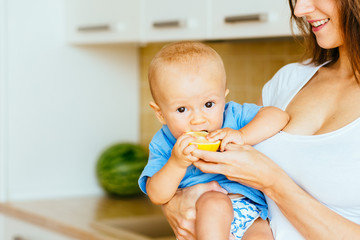 Portrait of cute healthy baby boy in blue shirt eating sour lemon on mother's hands at home kitchen interior.