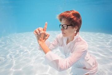 Portrait of young woman in office white shirt and glasses swim and pose underwater holding fingers in gun gesture. Businesswoman Diving in water pool, Dreaming of Holiday vacation time concept.