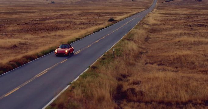 Aerial View Of Couple Driving Red Convertible Sports Car Down Country Road At Sunset 