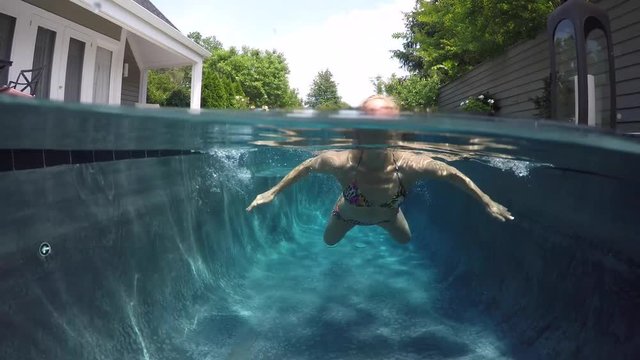 Camera half above and half below water, a beautiful, slim, caucasian woman wearing string bikini swims toward the camera.
