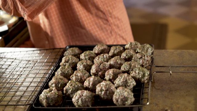 Man rolls balls of kofta in his hand and puts into grilling basket for cooking.  Camera slides from right to left.