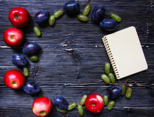 Apples, plums, grapes and notepad on a black wooden background