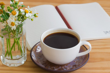 Black coffee and white notebook open on rustic wood table with brown or black pencil. Open notebook with pencil and vase of chamomile beside coffee. Copy space concept for background or wallpaper.