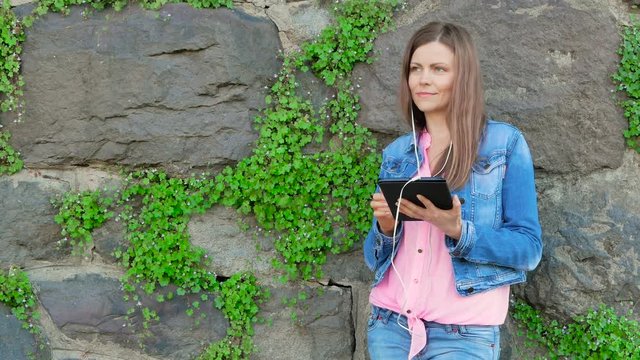 Pretty girl in a cotton jacket using tablet computer with earphones. Vintage wall of wild stone in the background.