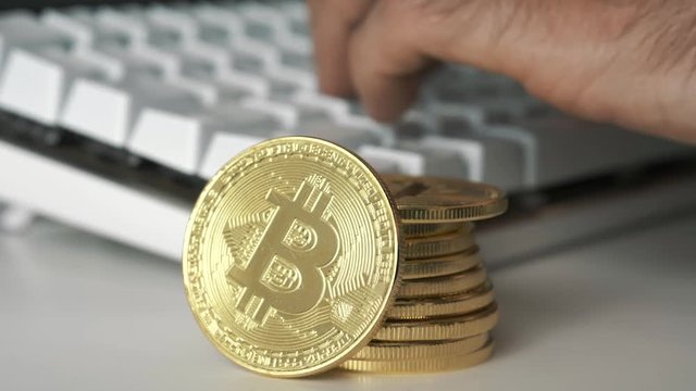 A stack of bitcoin coins while a hand is typing on a computer keyboard in the background. Bitcoin is a worldwide cryptocurrency and digital payment system.