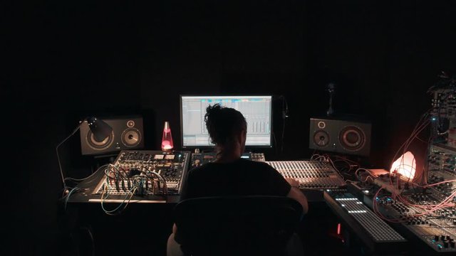 Young professional creative music or songwriter creates tunes for project, in dark room workstation studio uses computer software and recording devices, multiple cables and lights