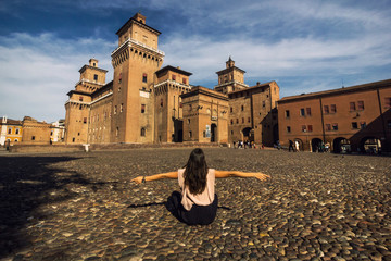 Girl sitting on a cobble-stone street to view Castello Estense of Renaissance town of Ferrara - obrazy, fototapety, plakaty