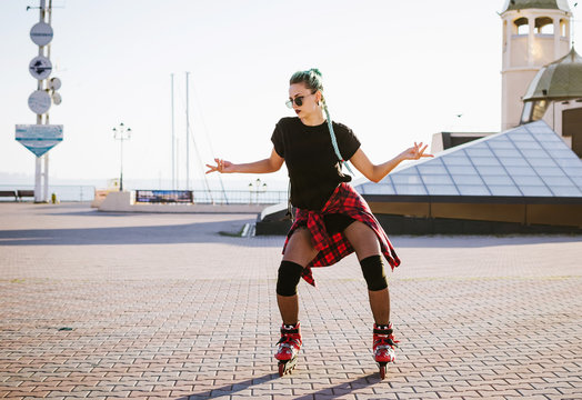 Young stylish funky girl with green hair riding roller skates and dancing near sea port during sunse