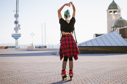 Young stylish funky girl with green hair riding roller skates and dancing near sea port during sunse
