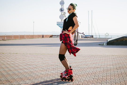 Young stylish funky girl with green hair riding roller skates and dancing near sea port during sunse