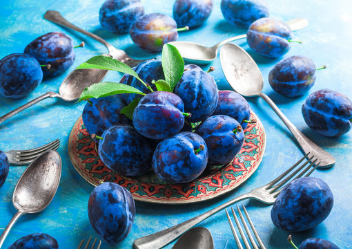 Colorful closeup plums group with leaves on plate and painted blue background with forks and spoons in studio