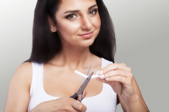 Stop Growing. Portrait Of A Beautiful Smiling Woman Holding Cigarettes And Scissors In Her Hands. The Concept Of Purchasing Cigarettes. On A Gray Background.