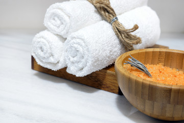 Orange bath salt in a wooden bowl and three white towels on a wooden stand stand on a white marble table
