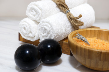 Orange bath salt in a wooden bowl and three white towels on a wooden stand and black Bian stones stand on a white marble table