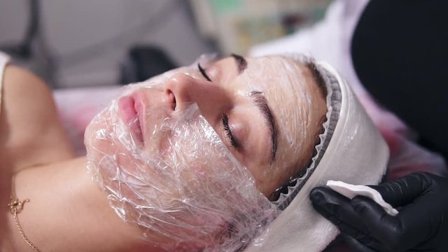Professional cosmetologist is cleaning woman's face from special treatment using cotton sponge. Young woman is lying on the couch during cosmetic face procedure in spa salon