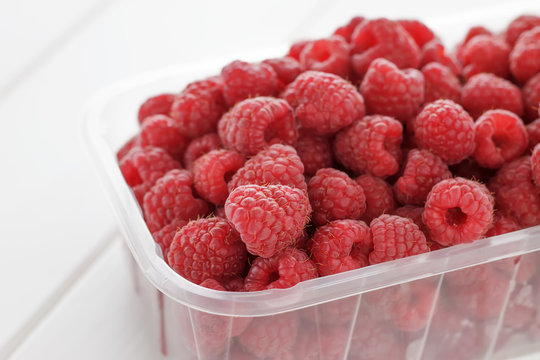 Fresh raspberries in plastic box from local market on white wooden background.