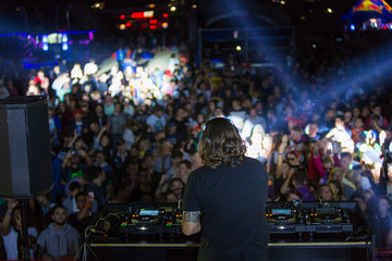 DJ with headphones at night club party under the colorful light and people crowd in background. Dj at open aire party. Night party at sea coast. Beach party.