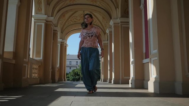 Beautiful girl in blue pants and pink lace top walking in historic architecture. Yoga in the city. Health concept. Slow motion. Woman goes to the camera and smiling.