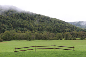 Hillside with low clouds and green meadow with fence section
