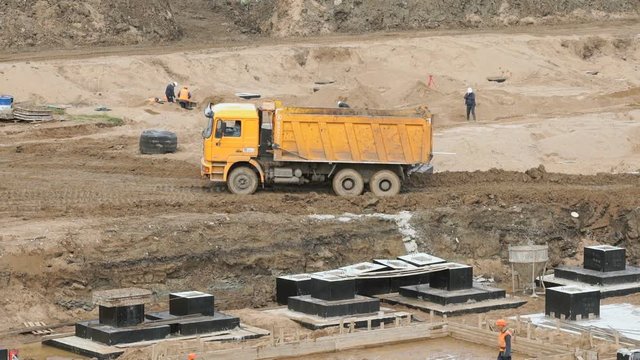 Yellow truck drives on the clay at the construction site in summer. Workers in helmets and suits level surface