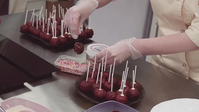 Unrecognizable confectioner in rubber gloves decorates chocolate ball on stick with colorful confetty at kitchen over metal desk