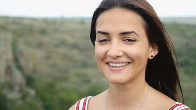 A cheerful and beautiful girl smiles happily atop some cliff on a sunny day in summer in slow motion. The landscape and cloudscape look inspiring and splendid