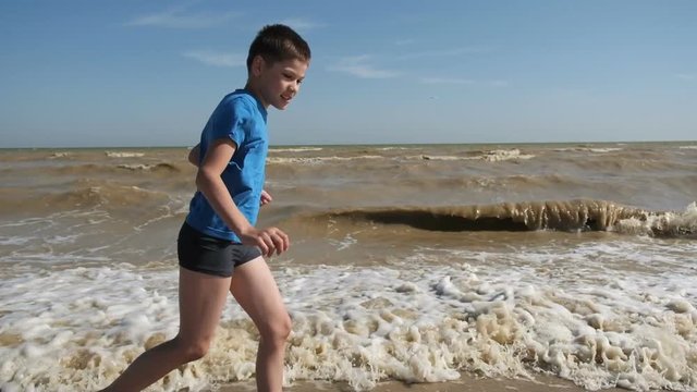Boy run along the beach. A playful young boy dashes along the seashore, with waves splashing around his feet, enjoying a sunny beach day. Slow motion