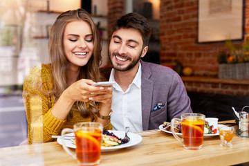 Cheerful young couple sitting together and eating breakfast in cafe taking photo of their food and smiling