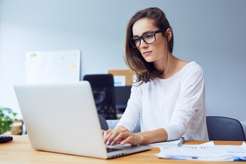 Serious beautiful young woman typing on laptop in a bright modern office - Powered by Adobe