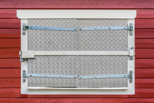 Closed window on red exterior wood wall with security fittings, bars, lock and metal shutters.