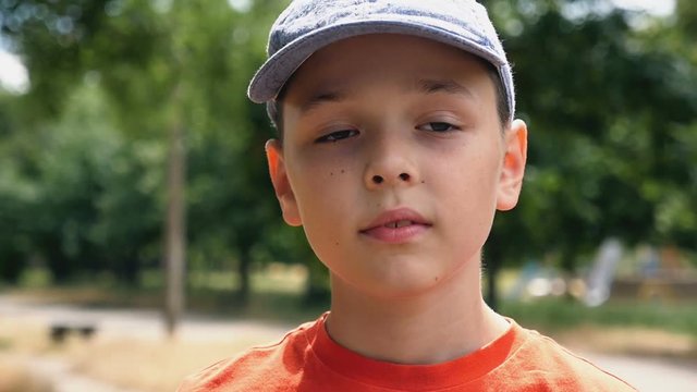 A kind seven-year-old boy with deep eyes and a cap on his head stands in a park on a picturesque lawn and thinks about his life with on a sunny day in summer in slow motion