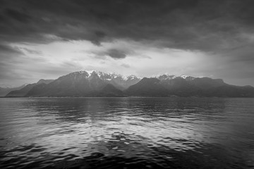 Snow on mountain peaks by the Lake Geneva in cloudy rainy weather in Lausanne, Switzerland. Black and white photography. Dramatic sky.