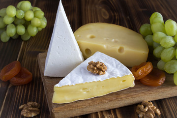 Different sorts of cheese, grapes and dried apricots on the brown cutting board