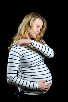 Young beautiful pregnant girl with blond curly hair  hugs her belly and touch her painful neck on black background, vertical image