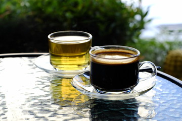 Black coffee in clear glass is placed on a glass table in front of a hot tea cup in a coffee shop.