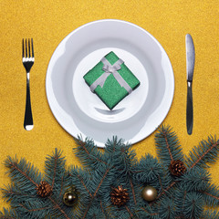 White plate with cutlery and gift box on a gold background. Branches of a Christmas tree with cones and toys. Happy New Year 2018. Christmas cooking. Holiday menu