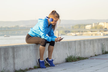 Young long hair girl with headphones on her head and phone on hand sitting and relaxing after jogging. The river and bridge is on background