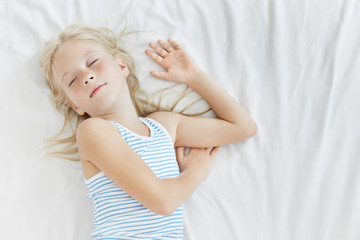 Candid shot of adorable Caucasian little girl with freckles wearing sailor pajamas while sleeping soundly in white bed, having daytime nap after first day at school. Innocent child napping in bedroom