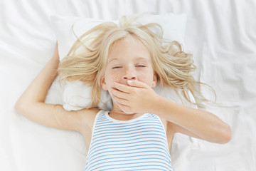 Sleepy pretty schoolgirl awakes early in morning, yawns, doesn`t want to go school. Exhausted freckled adorable child wants to sleep after hard day. Blonde attractive female kid has afternoon nap
