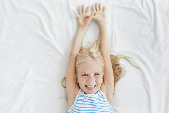 Bedtime Concept. Adorable Female Child With Freckles All Over Her Face Relaxing In Bed After Good Sleep, Stretching Arms Up And Sticking Out Her Tongue, Having Playful Look, Wearing Striped Pajamas