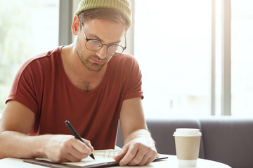 Young talented copywriter puts down ideas in notebook, sits at coffee shop, looks seriously at notes. Attractive hipster in stylish clothing being busy with writing, arranges meetings with people