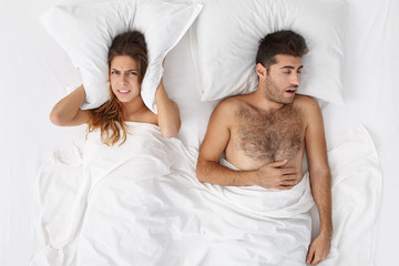 Frustrated stressed woman covering ears with pillow in bed while having sleepless night because of...