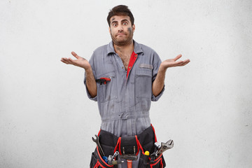 Studio shot of emotional clueless young unshaven male plumber with belt kit of working tools having...